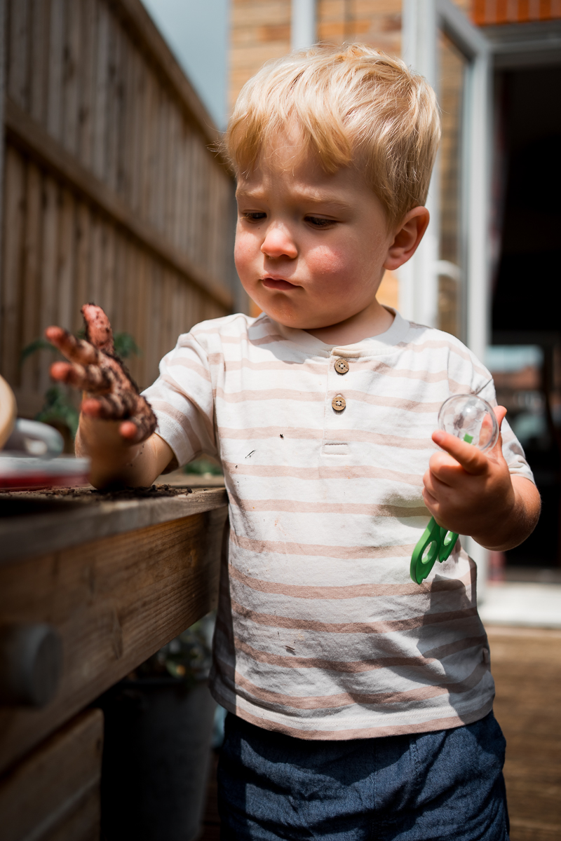 A toddler doing messy play outside looking at the mud in his hand.