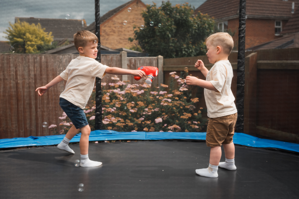 A young boy shooting soap bubbles on a toddler on a trampoline.