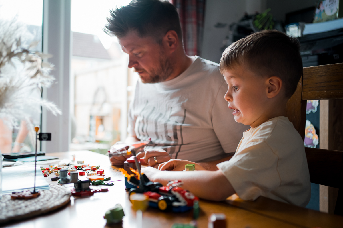 Father and son sitting it a table in front of a window playing with Lego.