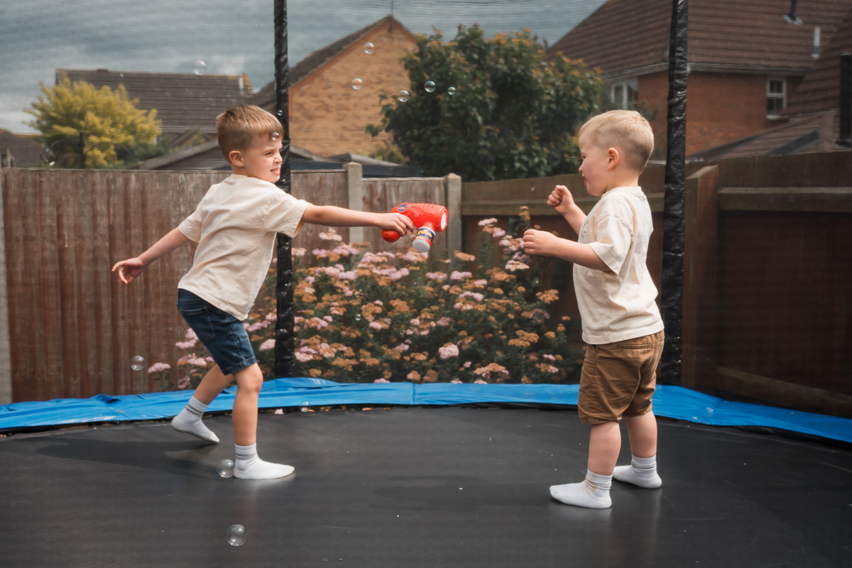 A young boy shooting soap bubbles on a toddler on a trampoline.