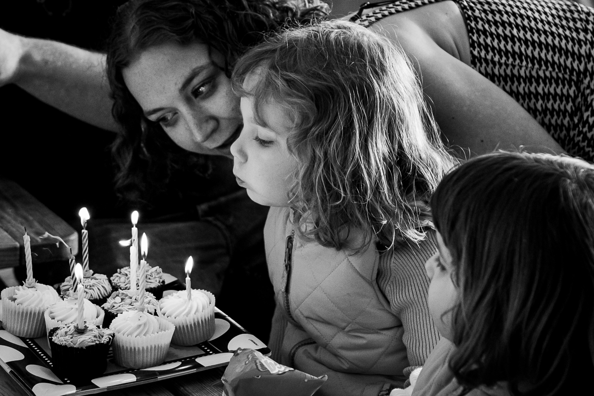 A young girl blowing out candles on birthday cupcakes surrounded by her mother and sister.